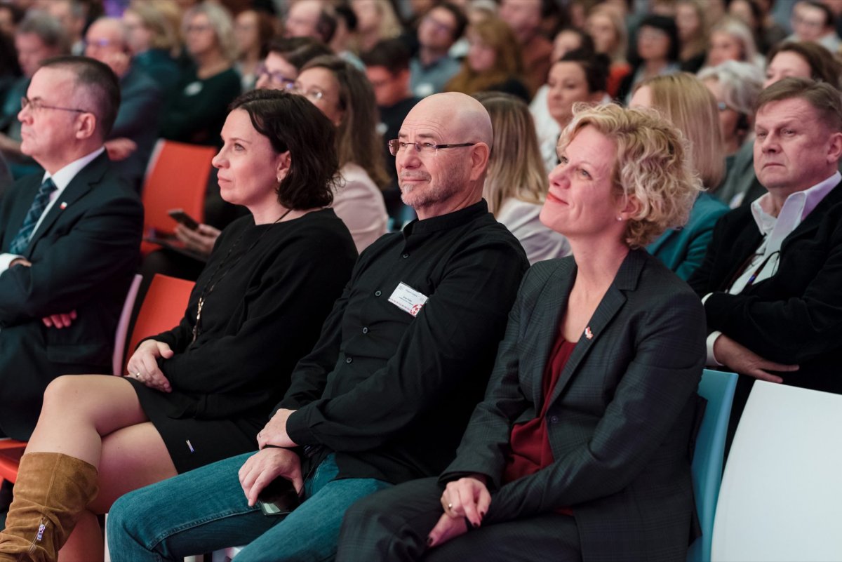 A group of people sit attentively in a large, crowded auditorium. Among them, a man in glasses and a black shirt is flanked by two women, one in a black dress and the other in a dark suit. The audience seems focused on an unseen speaker or presentation, captured elegantly by Marcin Krokowski's event photography.  