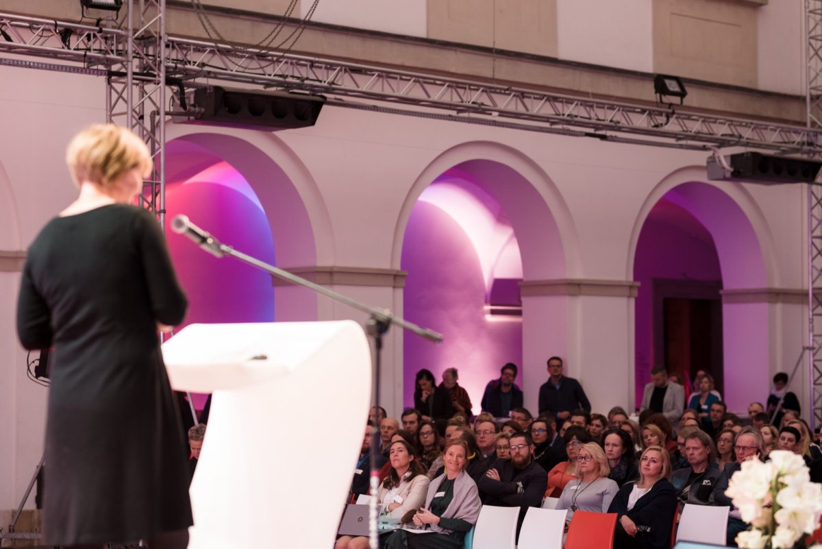 A person in black costume stands at a podium with a microphone and speaks to a seated audience in a large room with arched walls lit by colored lights. The audience listens attentively, with some standing at the back. Marcin Krokowski captures this exciting moment in his latest photo report of the events.  