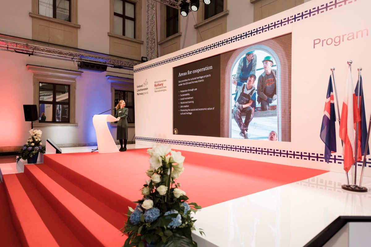 A woman stands on a stage lined with a red carpet decorated with white flower arrangements and gives a presentation. Behind her, a large screen displays images of employees under the heading "Areas of Cooperation." On the right side of the screen, a number of flags are visible, providing a fascinating photo-reportage of the events.  