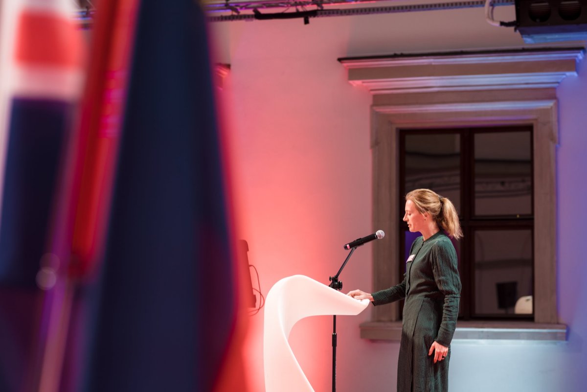 The woman stands on a white podium and speaks into a microphone in a room lit in red, blue and white. She is dressed in a dark green dress standing next to a window with an ornate frame. A piece of the flag is visible in the foreground, beautifully captured by Marcin Krokowski, a well-known Warsaw event photographer.  