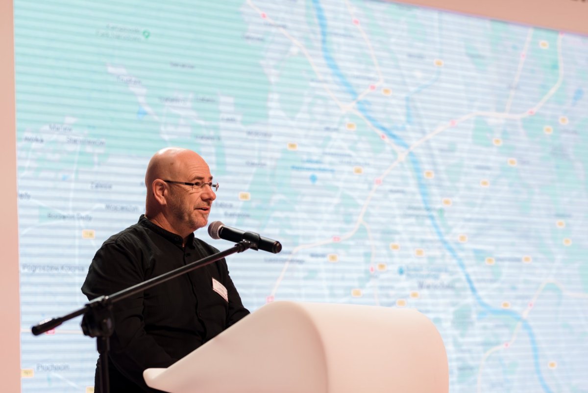 A bald man in a black shirt stands at a white podium and speaks into a microphone. Behind him, a map with various locations marked is projected on a screen. The event is captured by Marcin Krokowski, who specializes in photo coverage of the event.  