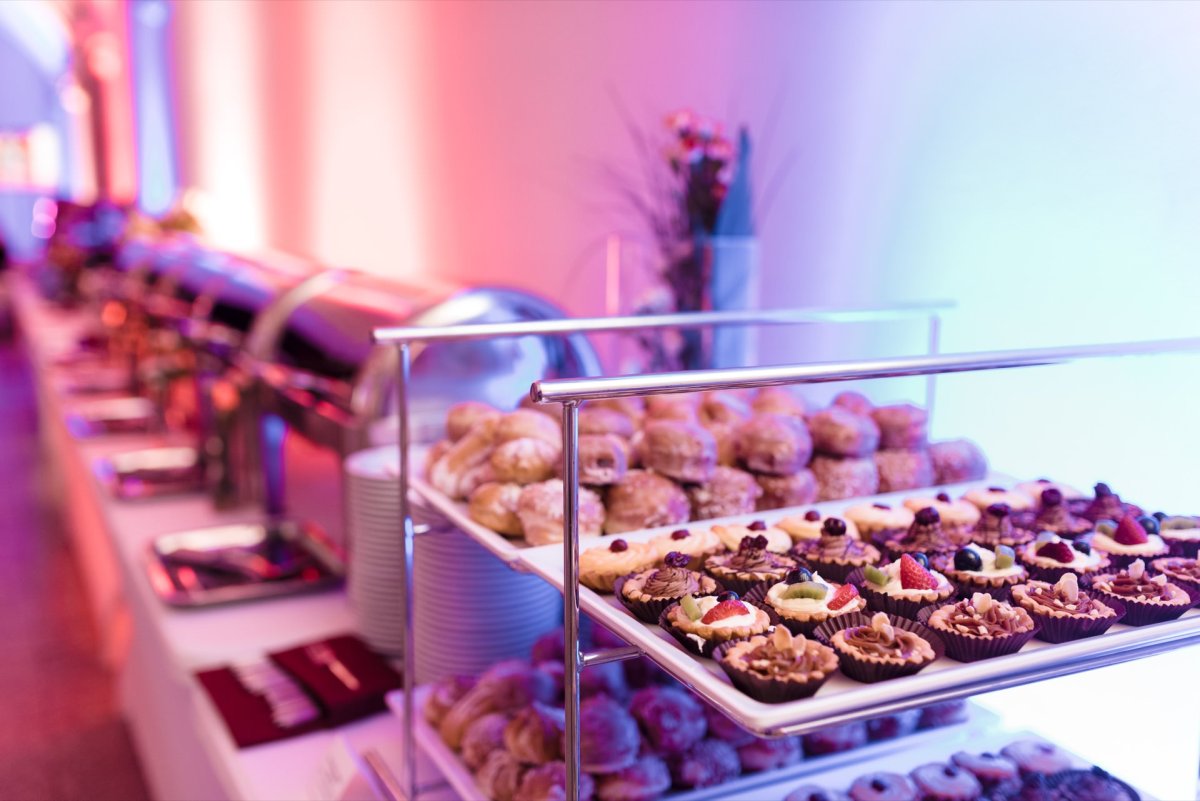 The buffet table is set with various trays of cakes and desserts, including mini fruit tarts. In the background are evaporation dishes with closed lids, and the table is illuminated by purple and blue ambient light. A perfect setting for event photography captured by Marcin Krokowski.  