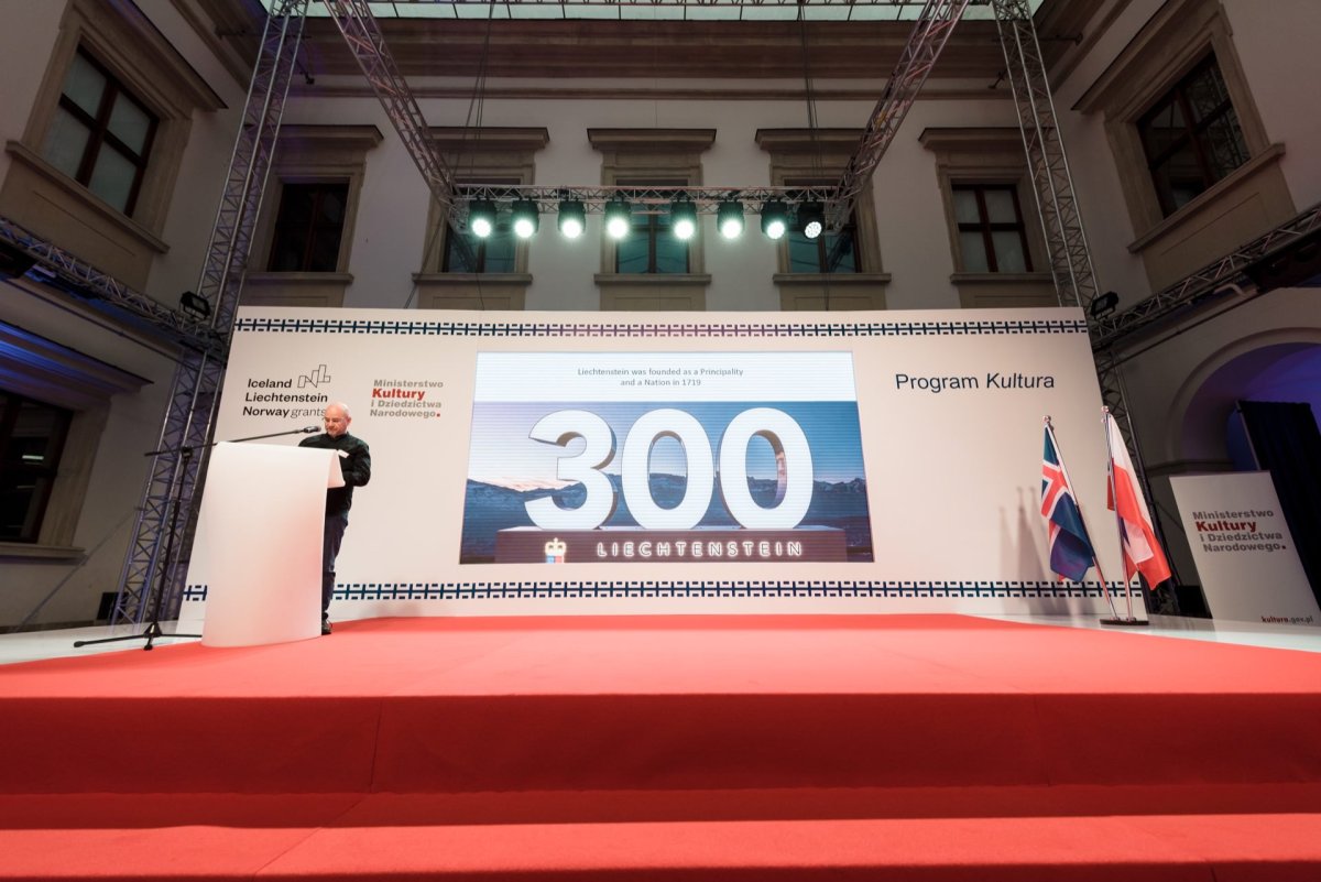 A person stands on a podium on a stage covered with a red carpet, and behind him is a large screen on which the number "300" is displayed in large numbers. The flags of Norway, Iceland and Poland are visible. This large room seems ideal for event photography or event photography by event photographer Warsaw.  