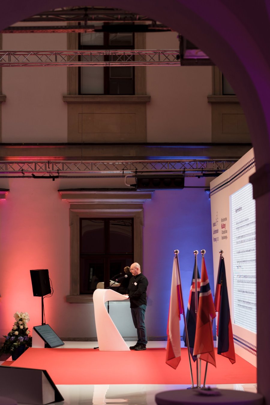 A person stands on a white podium, giving a speech in a large, elegantly lit room. Red and blue lights illuminate the stage, which displays several national flags, including those of Norway and Poland. A large screen displays the text to the right - the perfect moment for any event photographer Warsaw or photojournalism event.  