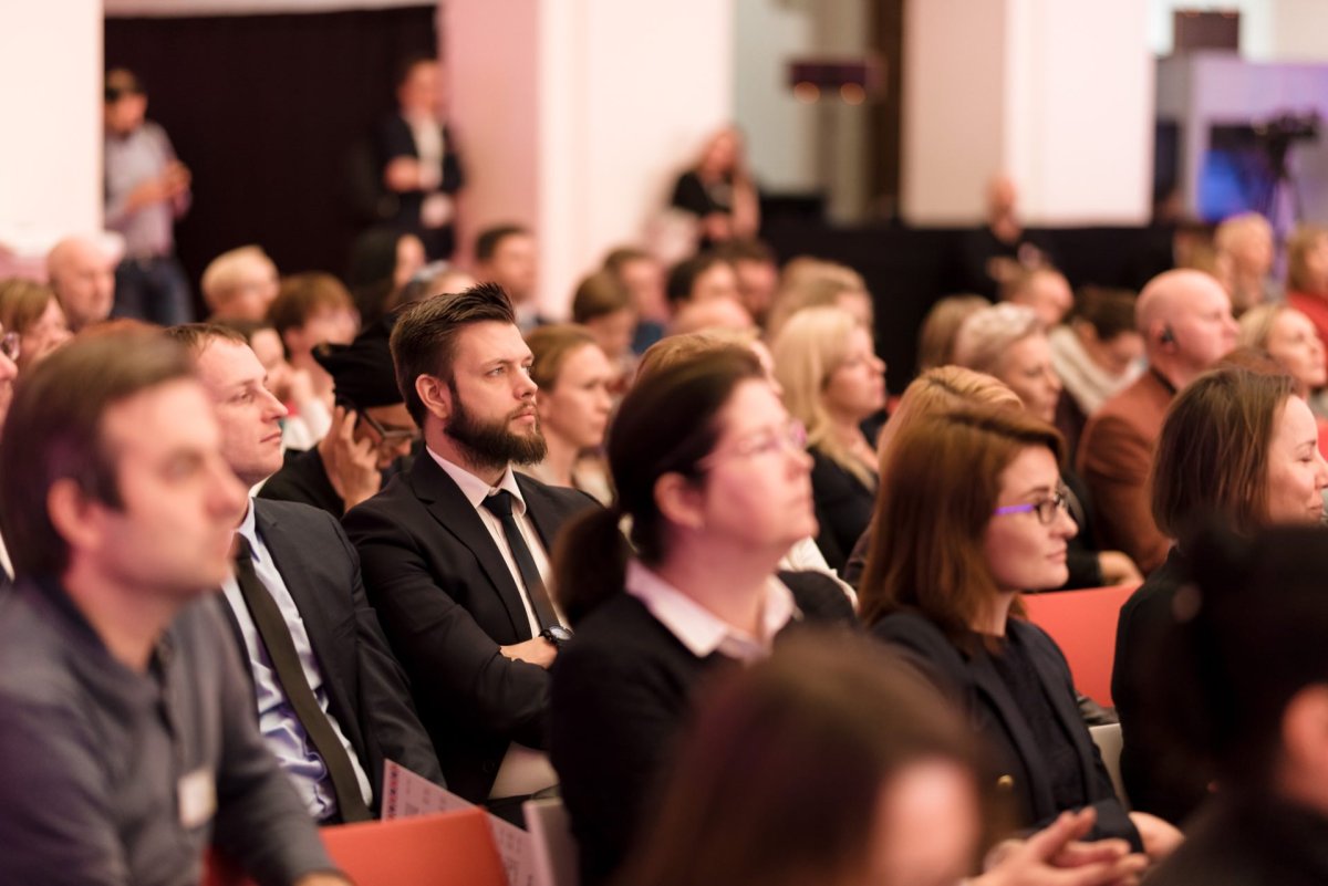 A group of people sit attentively in rows of chairs during what resembles a conference or seminar. Spectators in business attire face forward and listen to the speaker off-camera. The backdrop is a blurry interior with more people visible - captured in a detailed photo-report of the events.  