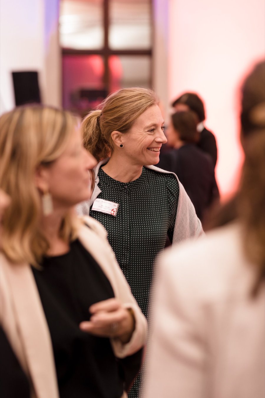 A woman wearing a green dress and gray sweater smiles while standing in a busy room. Surrounded by other people, the background is slightly blurred, indicating a photo report from an event or a social networking event. 