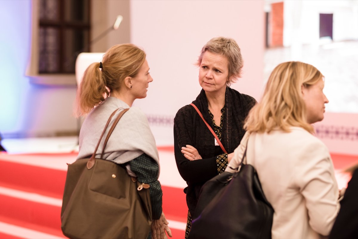 Three women are having a conversation at an event or conference in a room. One woman on the left is with her back turned; the middle woman is talking while looking at someone; and the right woman is slightly turned away. This scene captures the event's photo moment, highlighting the natural interactions.  