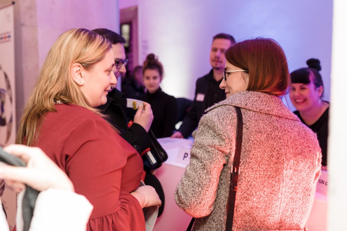 A group of people are having a conversation during an event held in a closed room. Two women in the foreground are facing each other and smiling. Several others are in the background talking at a table with name badges. The scene is warmly lit, perfectly captured by the skilled eye of Marcin Krokowski's photo report of the events.   