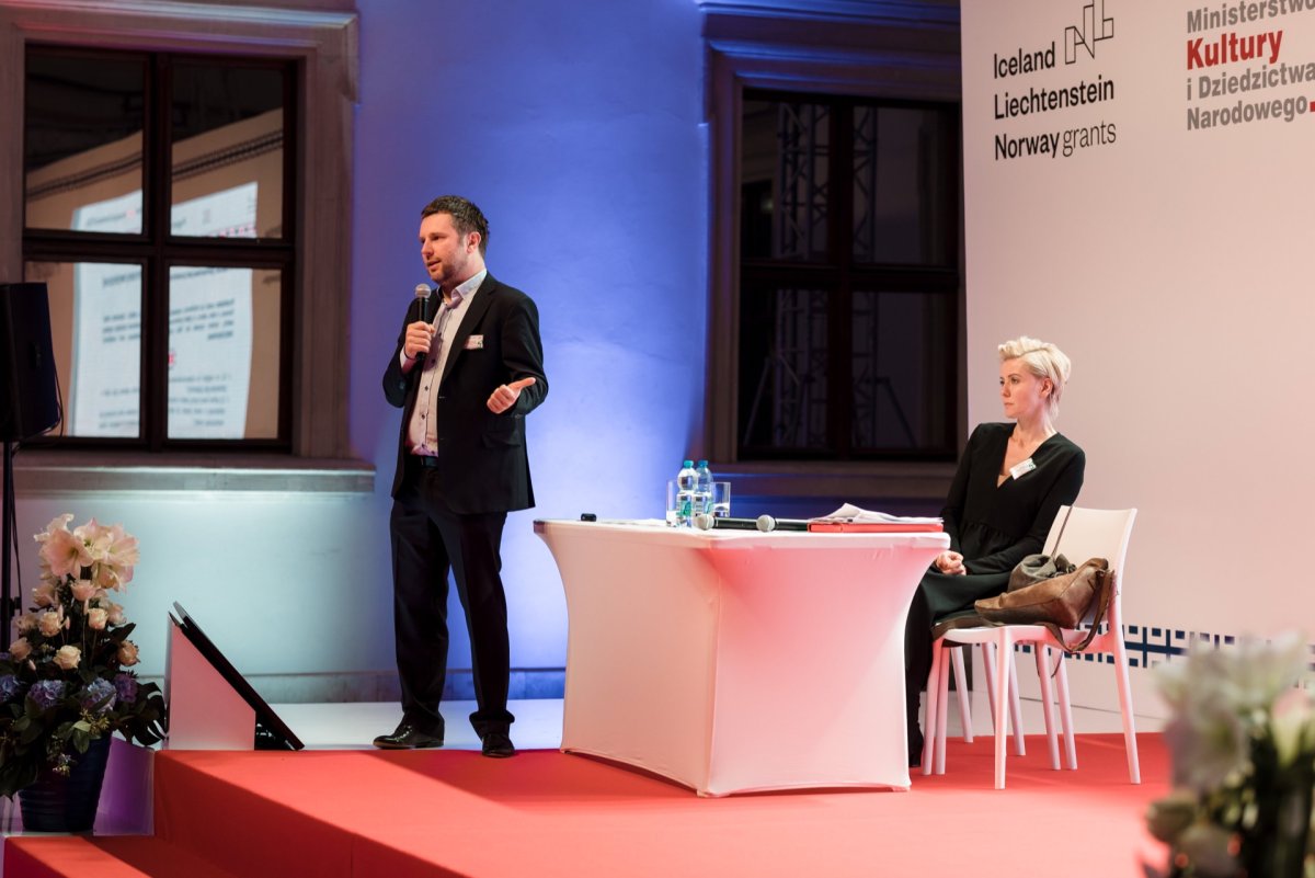 A man in a suit stands on a stage covered with a red carpet, holds a microphone and speaks. A woman with short blond hair sits at a white table with papers and a bottle of water and listens intently. In the background, text about the Iceland, Liechtenstein and Norway grant is displayed. This photo coverage of the event was captured by Marcin Krokowski.   