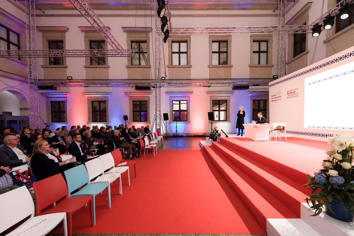 The speaker stands on a brightly lit stage next to a large projection screen in a large hall with high ceilings and large windows. The audience sits in rows of colorful chairs and listens intently. The room, perfectly captured in Marcin Krokowski's event photo report, is decorated with red and white flowers.  