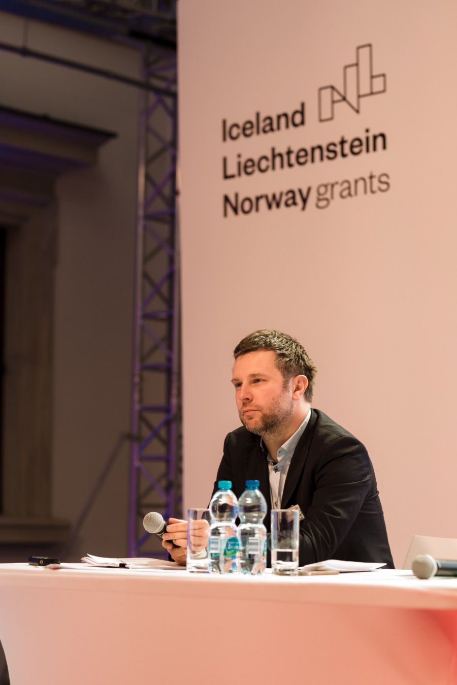 A man with short hair and a beard sits at the conference table, holding a microphone and a pen. He appears to be listening intently. In the background is the text "Iceland Liechtenstein - Norway Grants" along with a logo. Several water bottles stand on the table, capturing the essence of event photography.   