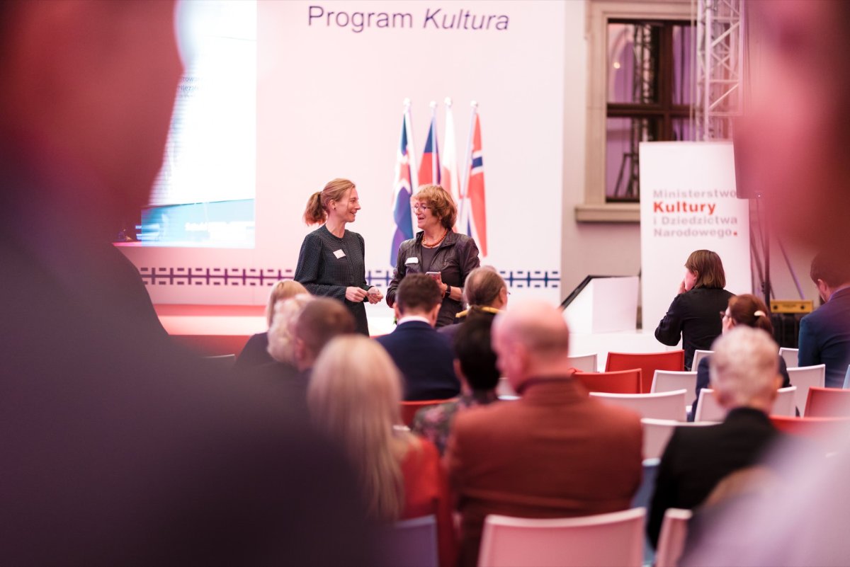 A group of people attending a formal event in a room. Two women are talking in the center of the picture. Behind them are flags, and a banner with the words "Culture Program" is displayed on the stage. A sign in the background displays the logo of the Ministry of Culture and National Heritage, captured by an event photographer Warsaw.   