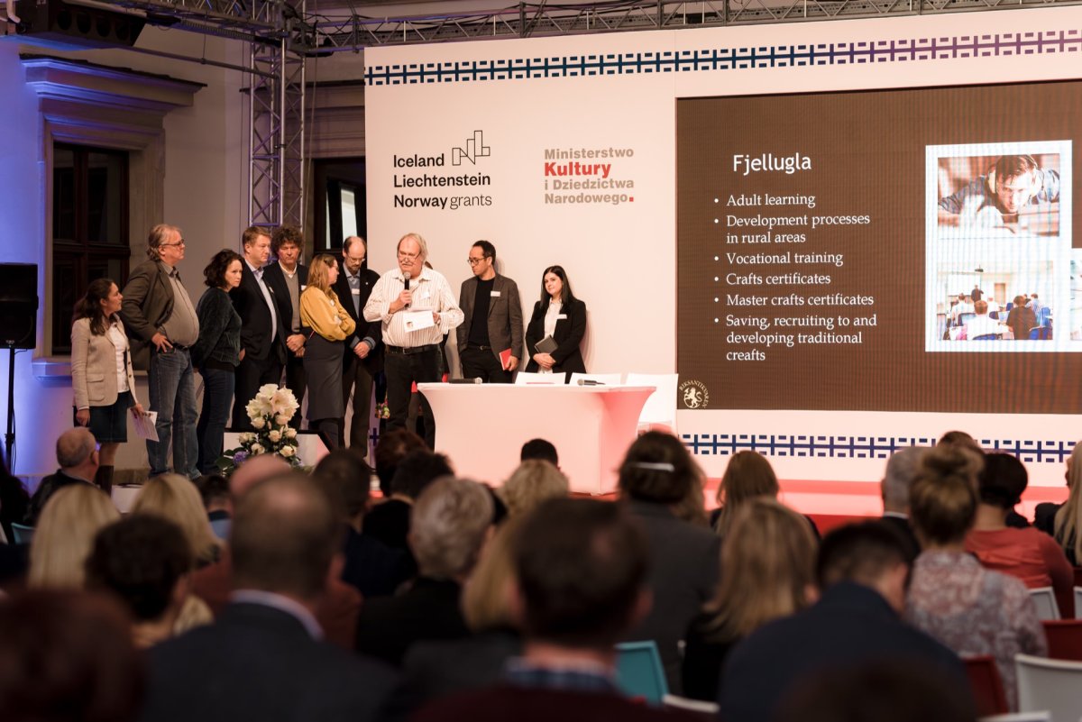 A group of people stand on a stage next to a large screen during a presentation at a conference. The screen shows the title "Fjellugla" and a list of topics related to adult education and crafts. In the foreground is the audience, who are intently watching the photo coverage of the event.  