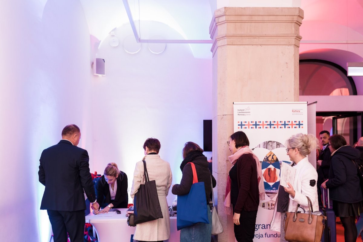 At the event held in the hall, people stand in line at the registration or information desk. The area is lit with soft purple and pink lights, creating an inviting atmosphere. They are assisted by a person behind the desk, and a banner with colorful graphics enlivens the setting, ideal for event photo coverage.  