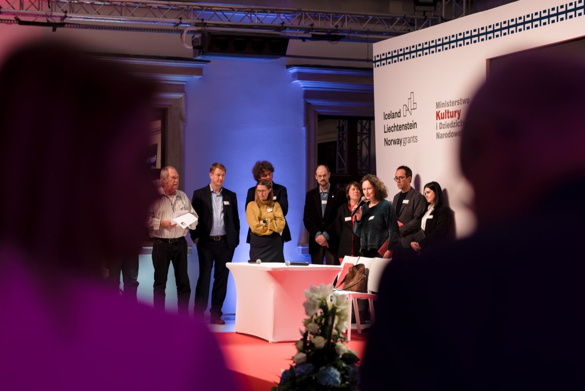 A group of people gather on the stage, some standing, others near the white podium. In the background is text on grants from Iceland, Liechtenstein and Norway. Captured by Marcin Krokowski, the stage is illuminated with colored light, creating a formal atmosphere for the event.  