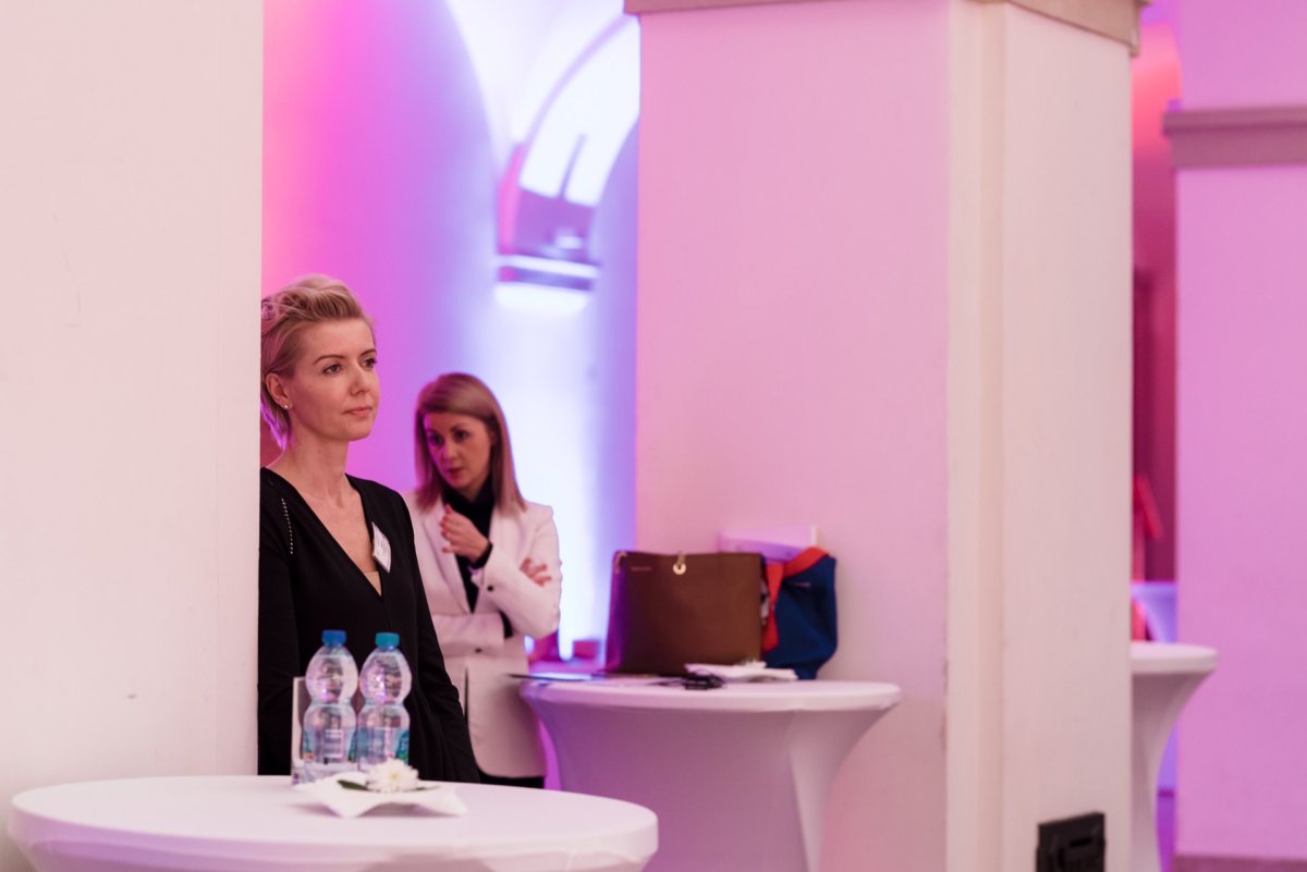 Two women are standing at cocktail tables in a softly lit room. The woman in the foreground leans against a pillar and looks thoughtful, while several bottles of water stand on a table in front of her. The woman in the background gestures as she talks; both are dressed in business attire, presenting moments from a photo shoot of a Marcin Krokowski event.  