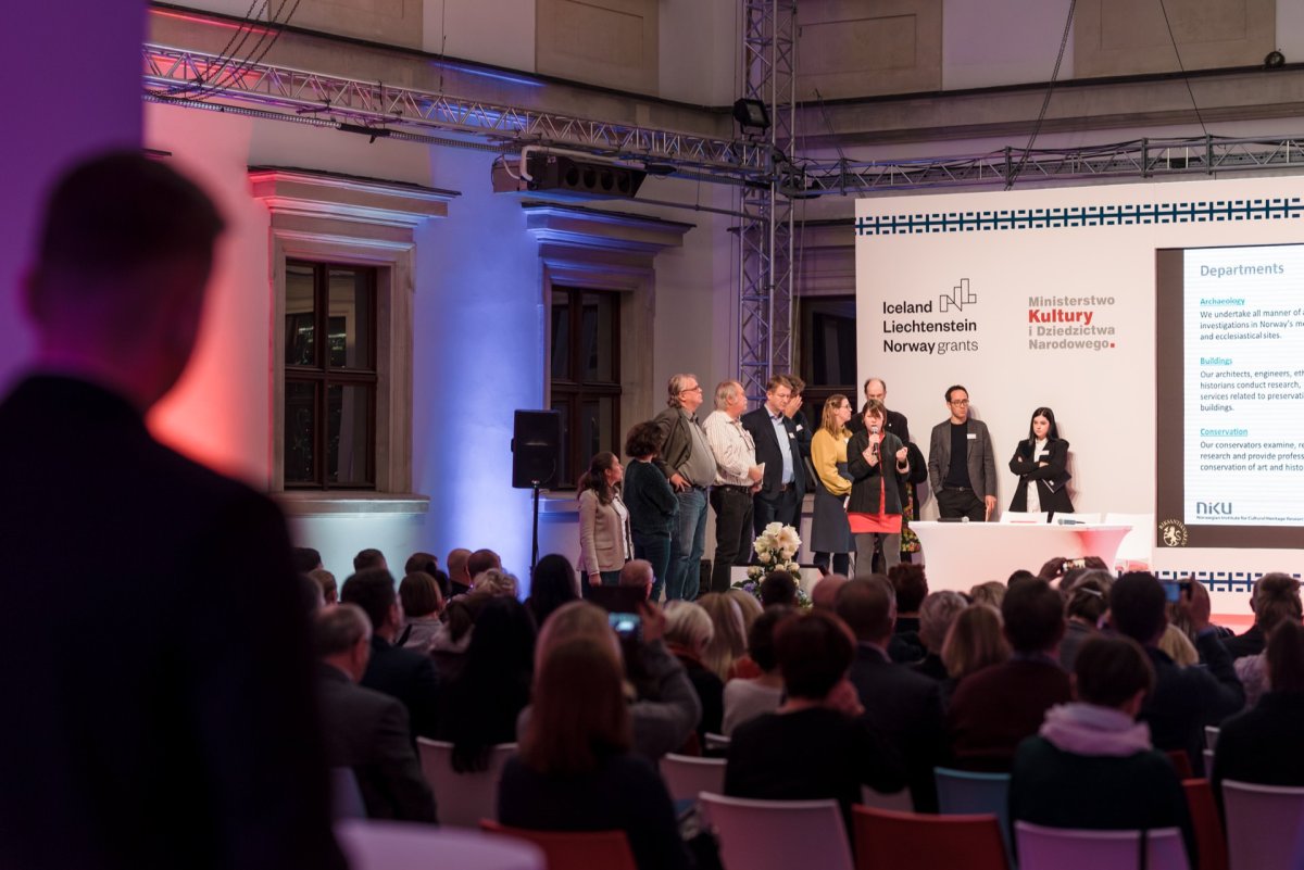 A group of people stand on a stage in front of a seated audience. A large screen displays text about departments and grants. The room is illuminated with colorful lights that perfectly capture the lively atmosphere, allowing any Warsaw event photographer like Marcin Krokowski to deliver an exceptional photo report of the event.  