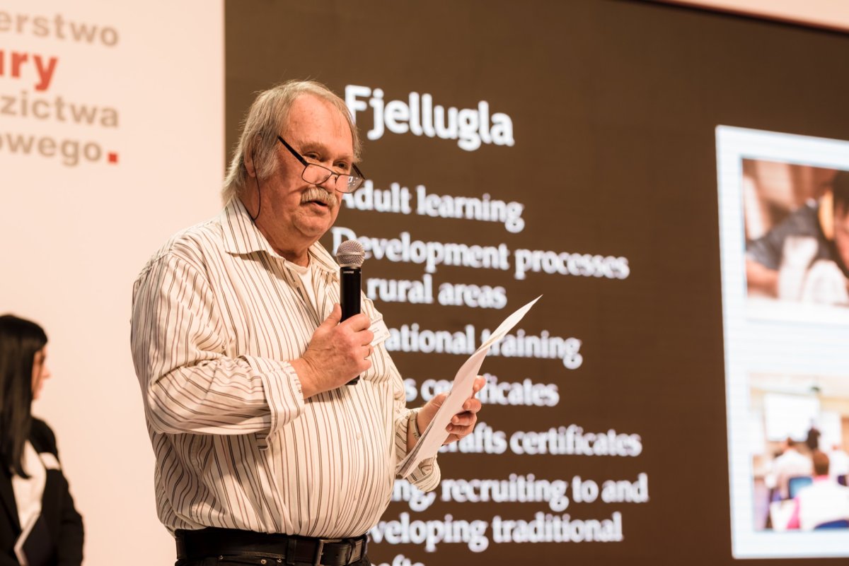 At the event, an elderly man with gray hair and a mustache speaks into a microphone while holding a newspaper. He stands in front of a screen displaying text about adult education and vocational training, ideal for photo coverage of events. The background contains text in various languages.  