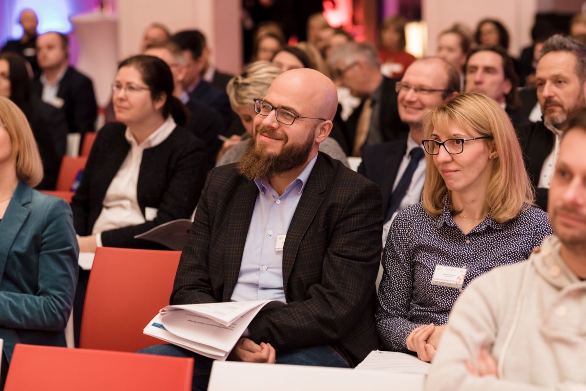 A group of people sit and listen attentively in a closed room. In the foreground, a bearded man in a checkered jacket and glasses sits next to a woman in a gray sweater and glasses. Both are smiling. In the background, you can see more people sitting in rows, perfectly capturing the atmosphere of the event photo shoot.   