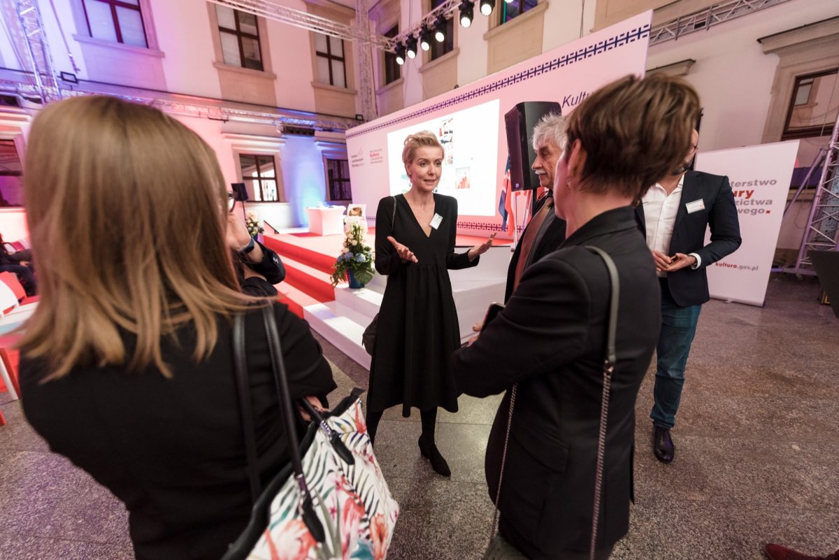 A woman in a black dress gestures as she talks to a small group of people during an event taking place in the room. The room is well lit with pink and white lights, complete with a staircase and floral decorations. People in the group listen attentively, capturing the essence of professional event photography.  