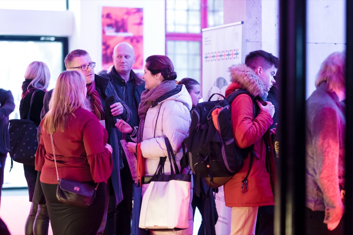 In the well-lit interior, a group of people dressed in winter clothes, including coats and scarves, are talking. Some are holding bags and carrying backpacks. In the background are fuzzy posters and lights, capturing the essence of the photojournalism of the event.  