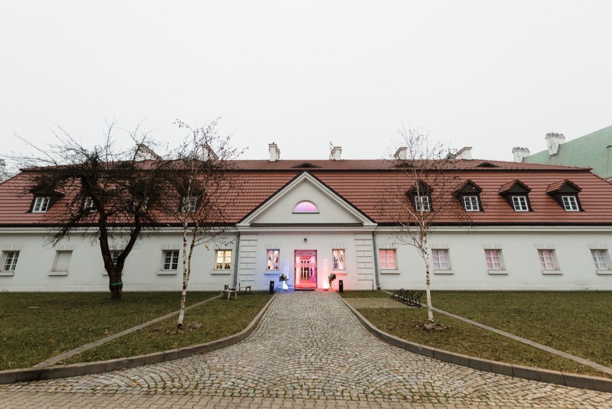 A charming white building with a red roof and dormers stands at the end of a cobblestone path surrounded by trees and grass. The entrance, lit with pink and blue lights, creates a welcoming atmosphere, perfect for the events captured in Marcin Krokowski's event photography. Higher up hangs an overcast sky.  