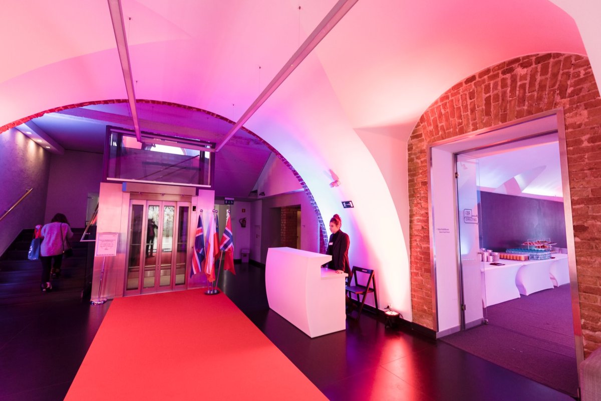 The modern, warmly lit lobby with arched brick walls features a reception desk, elevators and a red carpet. A woman stands behind a desk. To the right is visible a room with tables set up for the event. The colorful lighting creates a welcoming atmosphere, perfect for *event photography* occasions.   