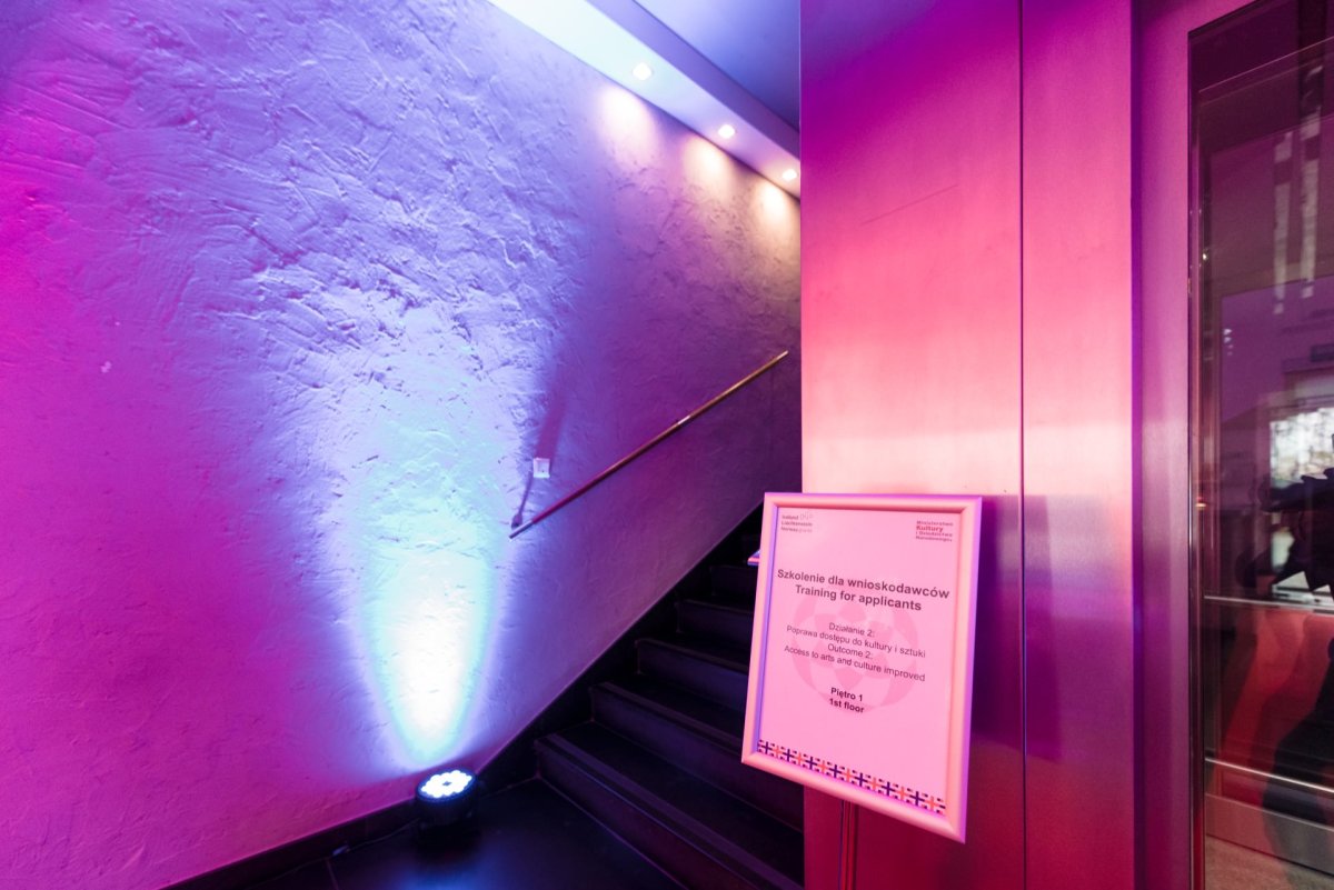 A sign at the entrance to the staircase with backlit purple and blue lights creates a striking atmosphere. The wall has a textured finish, and a handrail runs along the staircase. A reflective surface is on the right side. The sign seems to carry information, but the text is illegible, like a scene of Marcin Krokowski at an event photographer in Warsaw.   