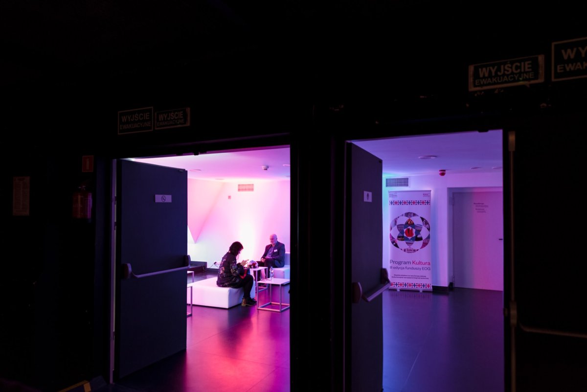 A dimly lit room with a glowing pink and purple atmosphere, visible through an open door. Two people sit on white furniture and carry on a conversation. Banners and signs with text adorn the space, and green exit signs glow above the door in the dark corridor outside - the perfect place for a captivating photo essay of the event.  