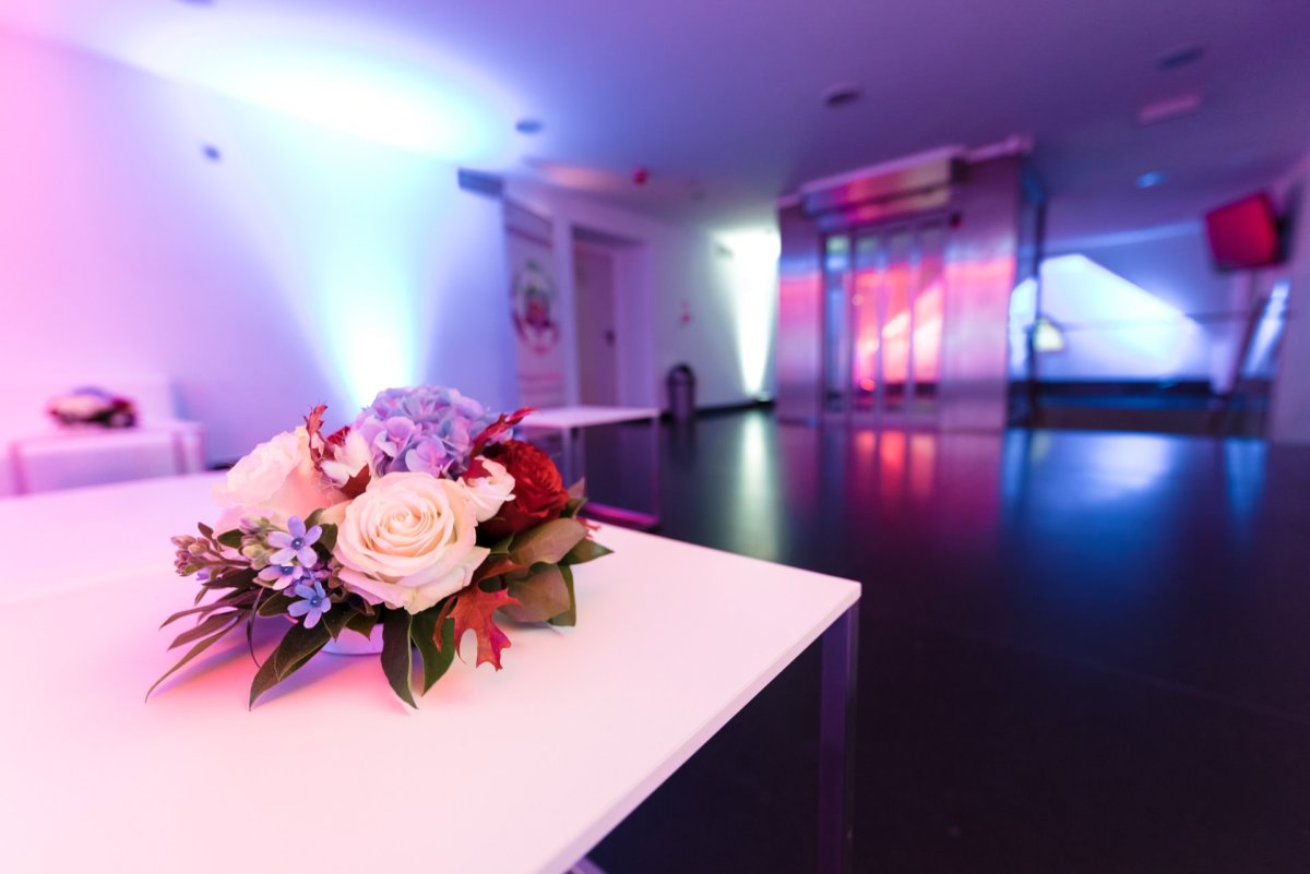 A small floral arrangement with white roses, blue and red flowers and green leaves sits on a white table in a modern room with vibrant purple, blue and pink lighting. The room has a sleek, minimalist design with an elevator and open space in the background - perfect for event photography. 