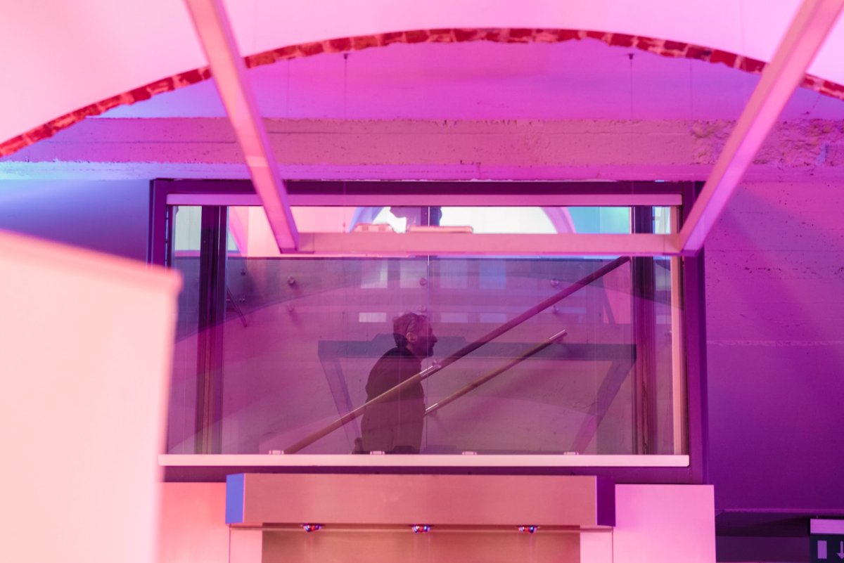 A person walks up the stairs into a modern building with purple and pink ambient lighting. Large glass panels and exposed concrete walls create a futuristic atmosphere, perfect for event photography or photo coverage. 