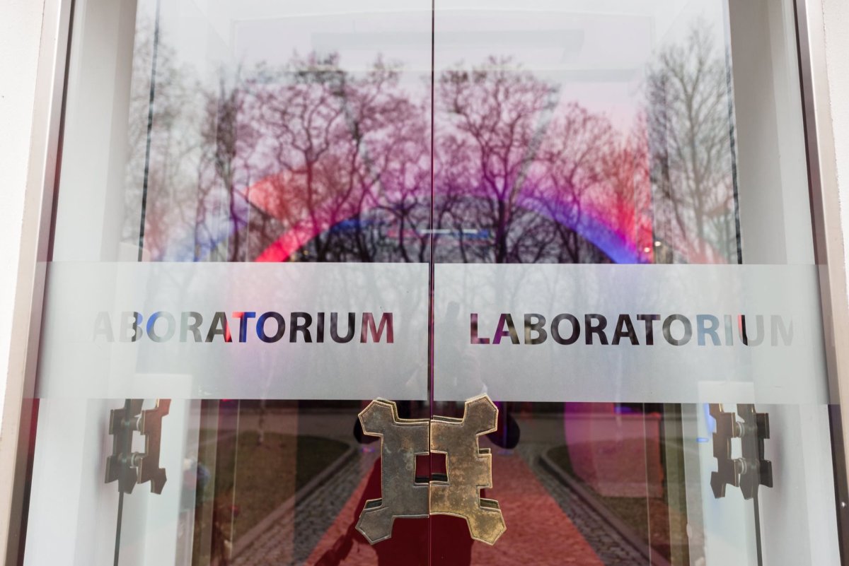 Glass doors with reflective surfaces show a blurred outdoor scene with trees and a path. The door features the inscription "LABORATORY" in black and colored letters and a metal handle in the shape of a stylized letter "H," reminiscent of the entrance captured in the event's photo gallery. 