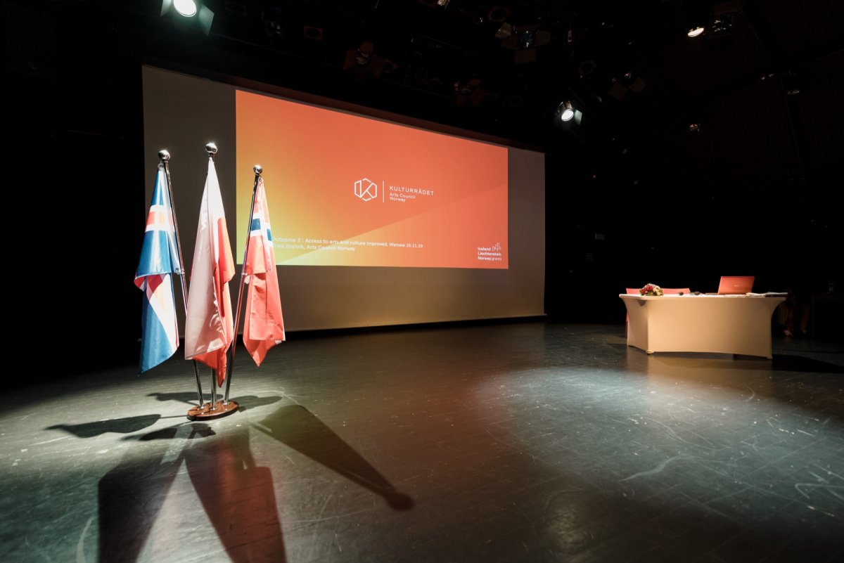 The dimly lit stage features a large screen displaying text and graphics, three flags on stands, and a white desk with a laptop and documents. The atmosphere suggests a formal setting for an event or presentation, perfect for capturing in event photography by well-known photographer Marcin Krokowski of Warsaw. 