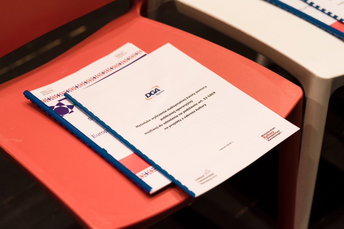 A document with the "DGA" logo lies on a red chair. The document looks like an official document with text in Polish. Next to it are other documents along with a binder and a graphic with the European Union flag motif. The chair is part of a row of seats, capturing a photo moment of the events.   