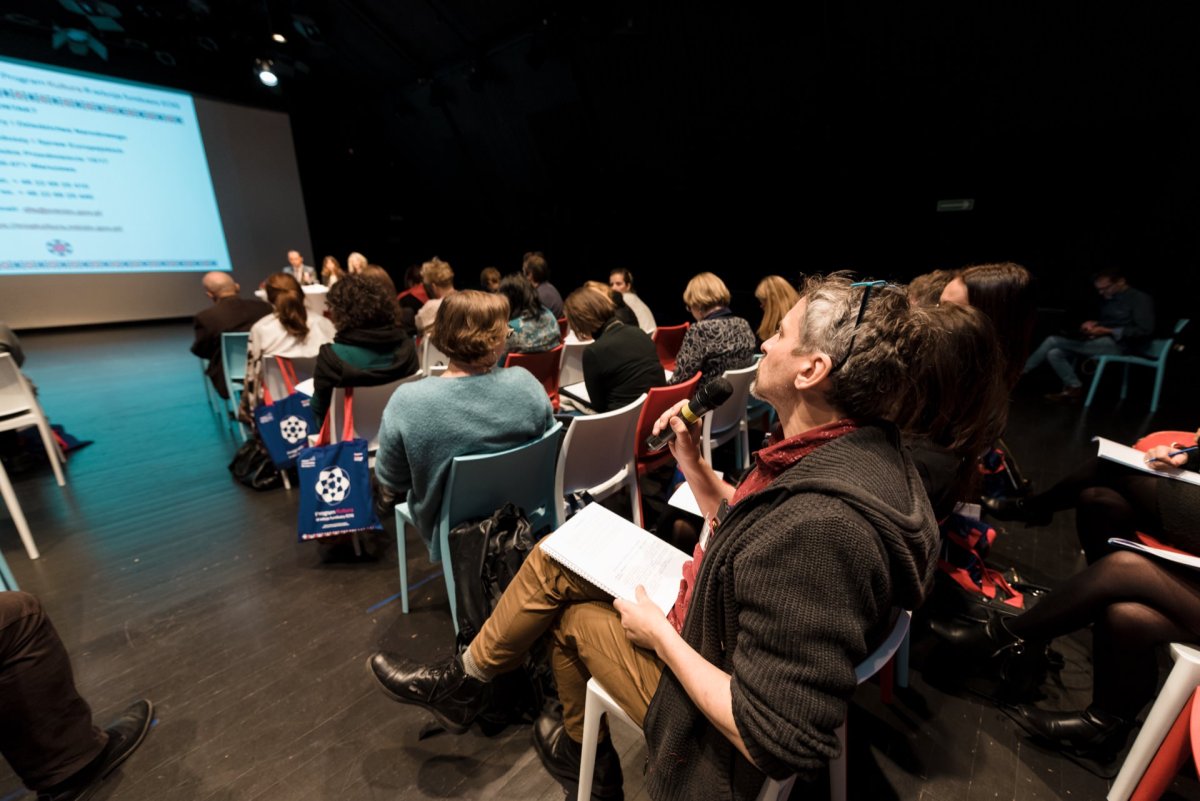 A group of people sitting in a dimly lit auditorium, facing a large screen on which a presentation is being projected. A person in the foreground holds a microphone and appears to be asking a question. Some participants have bags on the floor next to their chairs, capturing moments for a photo essay of the event.  