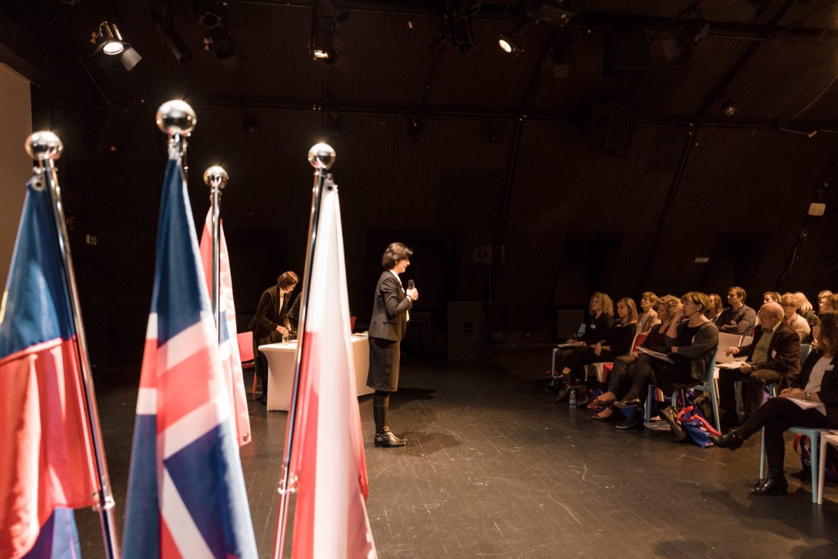 A speaker stands on stage and speaks to an attentive audience sitting in rows. Many flags are visible in the foreground, including a Union Jack flag. The scene appears to take place in a dimly lit auditorium or conference room, which was beautifully captured by event photographer Marcin Krokowski.  