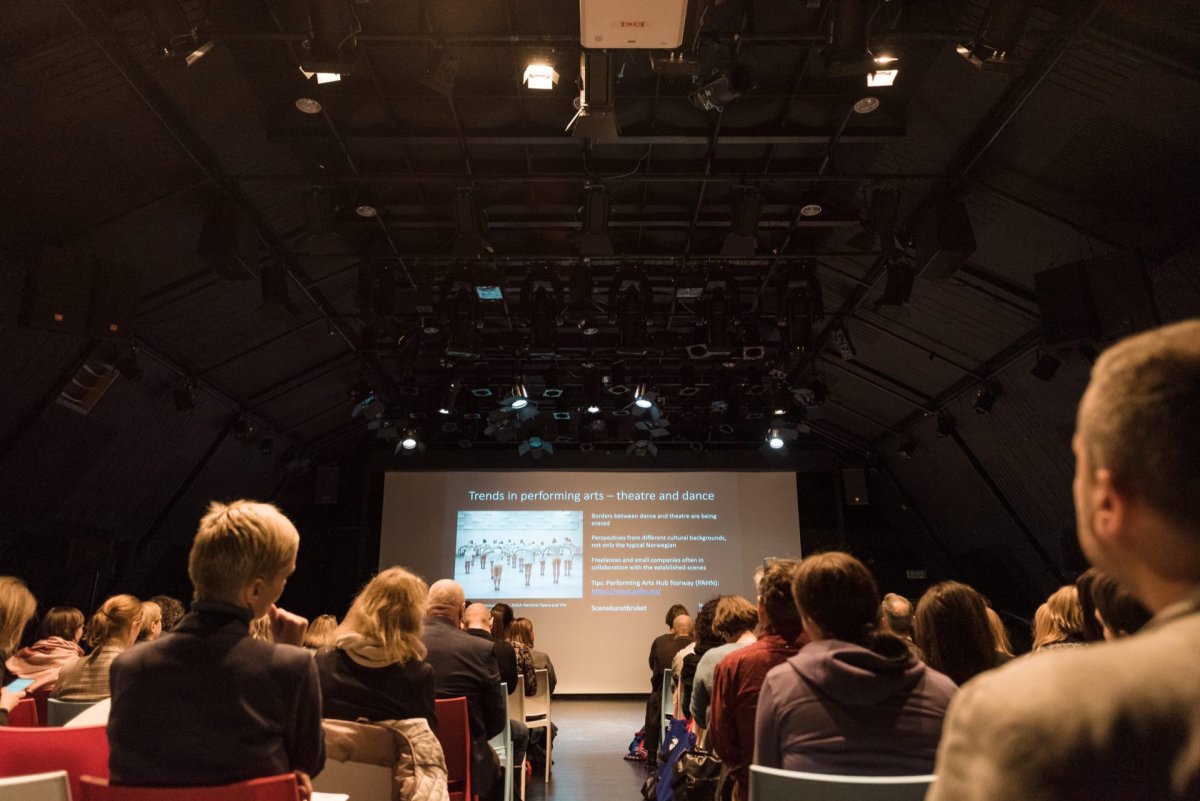 A large group of people sitting in the audience, facing a presentation screen that displays a slide titled "Trends in Performing Arts - Theater and Dance." The dark room is filled with a variety of equipment on the ceiling, and both men and women attend the event captured by event photographer Marcin Krokowski. 