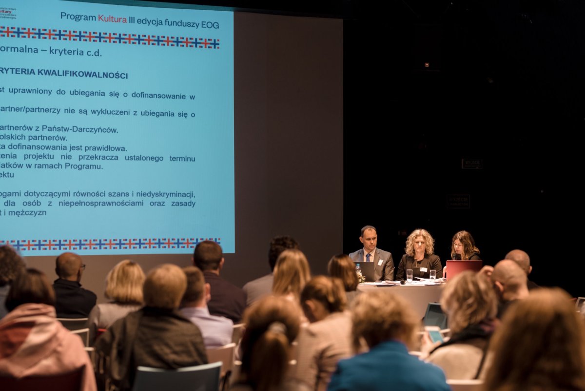 A conference room filled with seated attendees faces a panel of four speakers at a table on a stage. Behind them is a large projected slide with text in Polish and framed decorations. The room is dimly lit, focused on Marcin Krokowski's presentation.  
