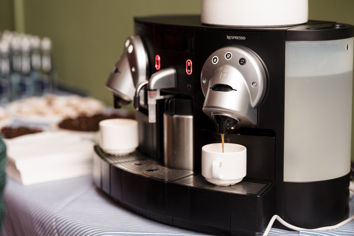 A Nespresso machine brews coffee into a white cup. Another cup stands next to it, presumably waiting to be filled. The machine stands on a table covered with a blue-striped tablecloth, and in the background you can see fuzzy objects, as if captured in one of Marcin Krokowski's photo essays of the event.  