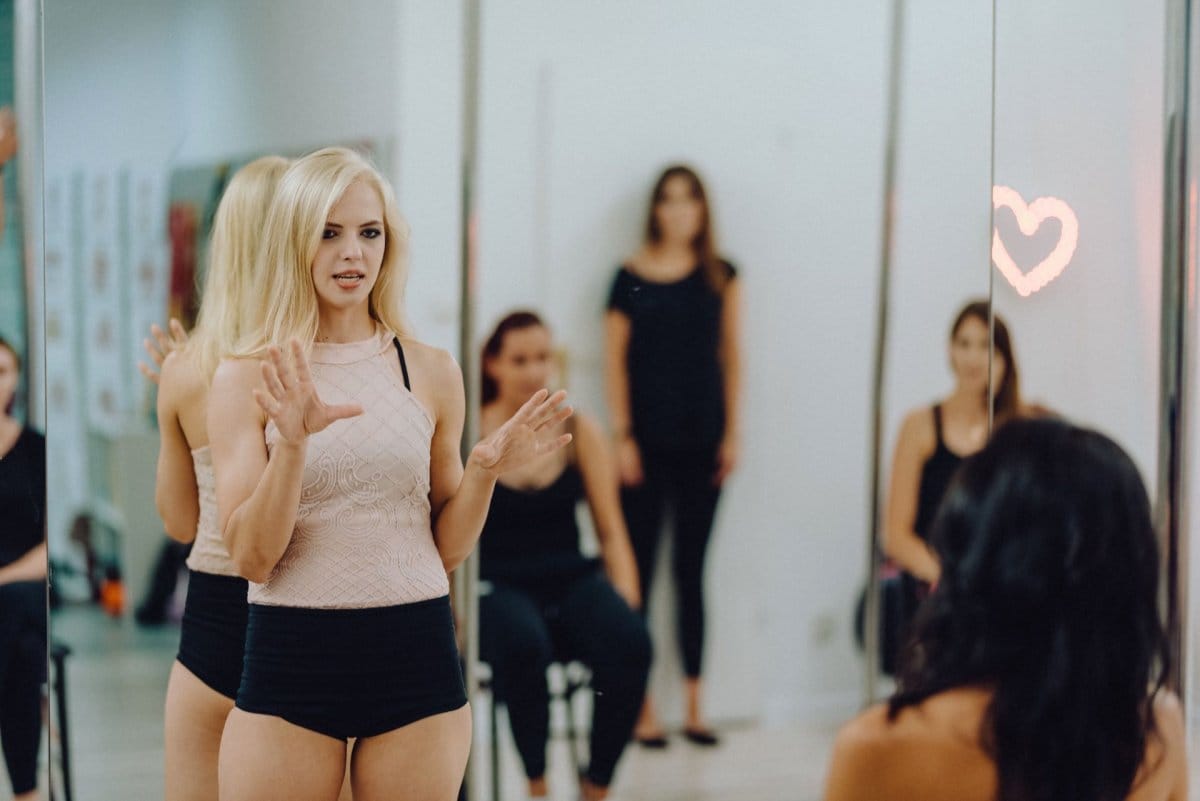 Blonde woman standing in front of mirror explains to girls in dance hall what to do