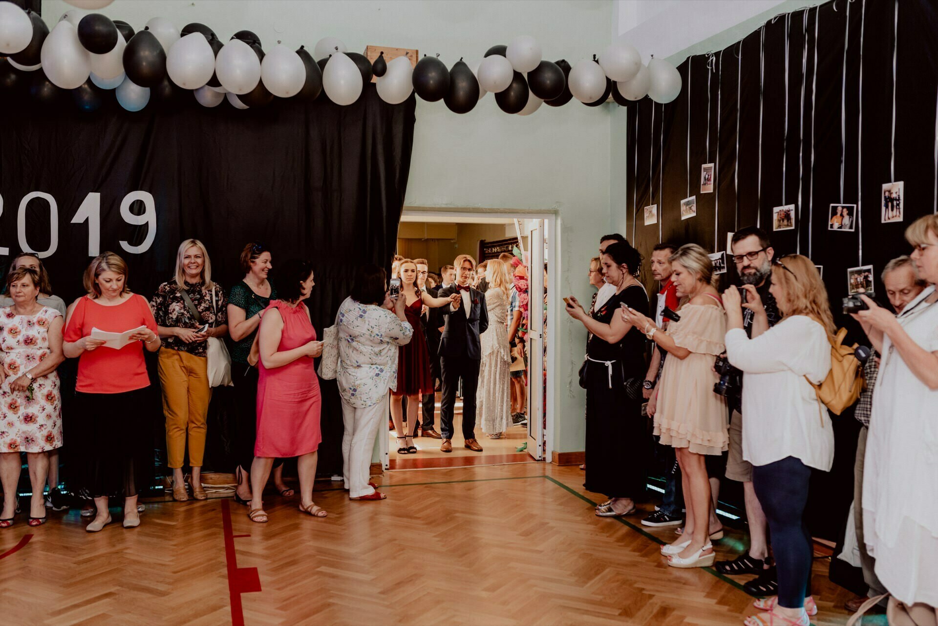 A group of people stand or lean against a wall under black and white balloons at the 2019 event, captured in this event photo. Some are clapping, others are using their phones. A figure in the doorway appears to be entering or exiting. The photos, likely part of an event photo tour, hang on strings along the right wall.