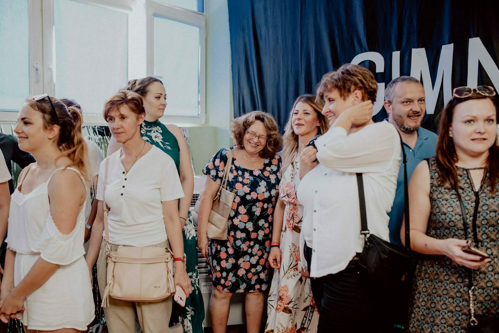 A group of people stand together in a room with natural light streaming in through a window. Some are smiling and talking, others are looking in different directions. The partially dark banner in the background, which shows the letters "GIMN," perfectly captures the moment as part of the event photography.  