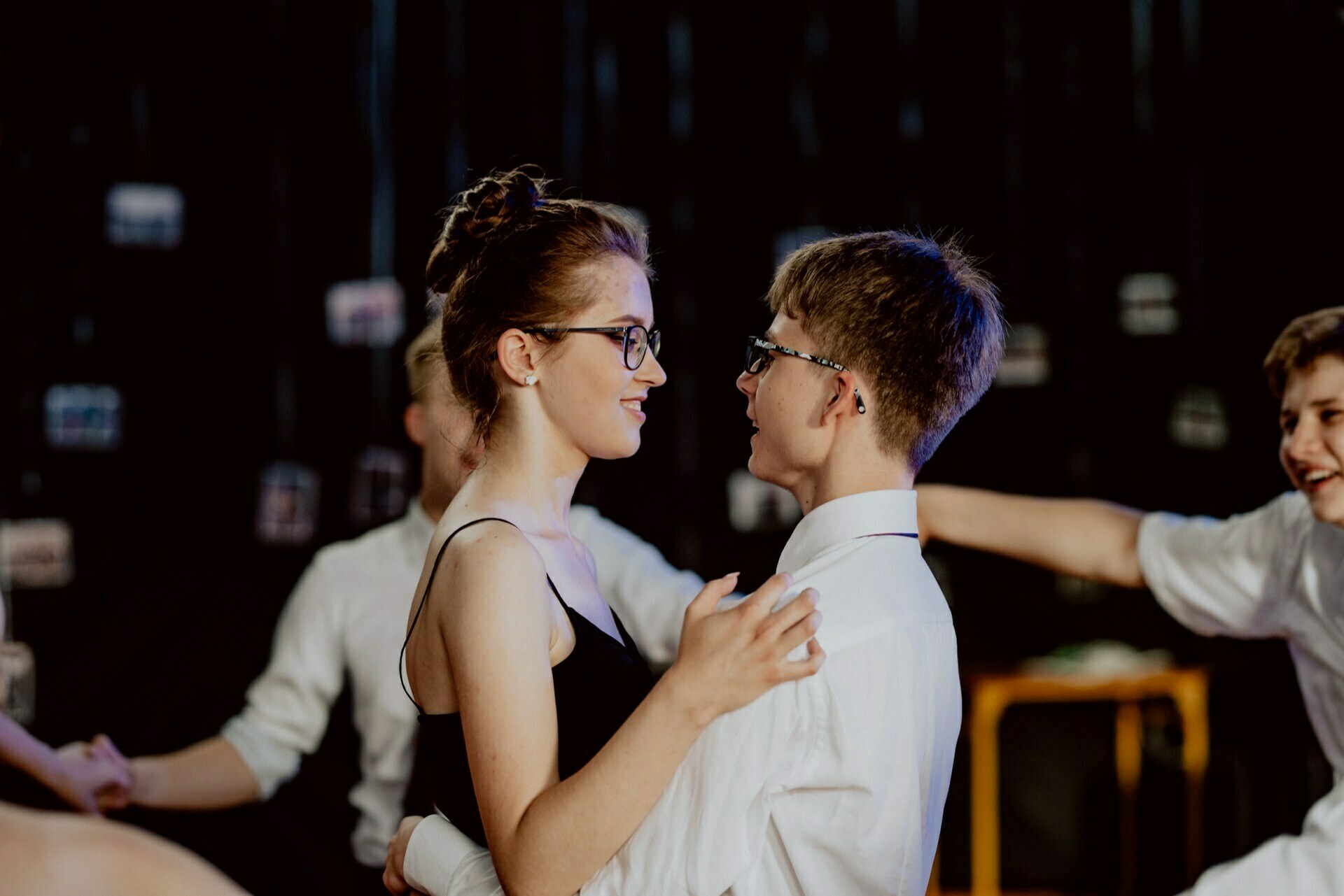 In a dimly lit dance hall, a young couple in glasses - a woman in a black dress and a man in a white shirt - dance close together, smiling at each other. Another couple moves in the background. This moment is perfectly captured as part of the event photographer Warsaw's event photo report.  