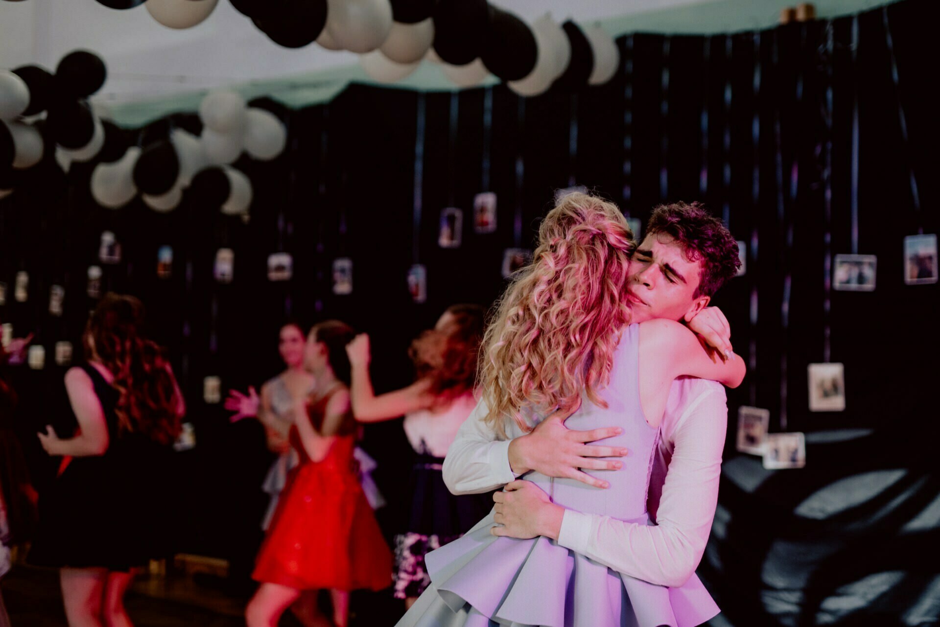 A young couple dances close, hugging during a formal event. The girl has blond curly hair and the boy has short dark hair. The partygoers are in the background, and the ceiling is decorated with black and white balloons. Photos hang on strings along the walls in this event photo.   