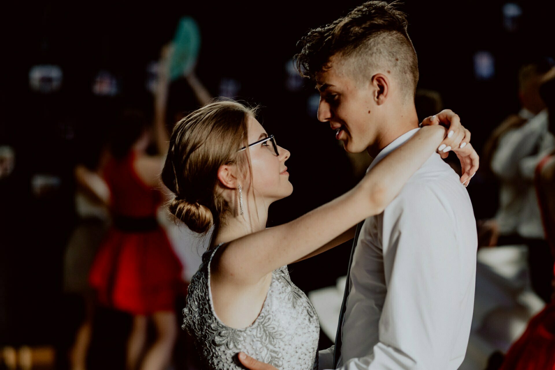 A young couple dances close, looking into each other's eyes. A woman in glasses and a silver dress and a man in a white shirt and short hair are the focal point of this beautifully captured event photograph. The background is softly lit and other dancing figures are blurred in the background.  