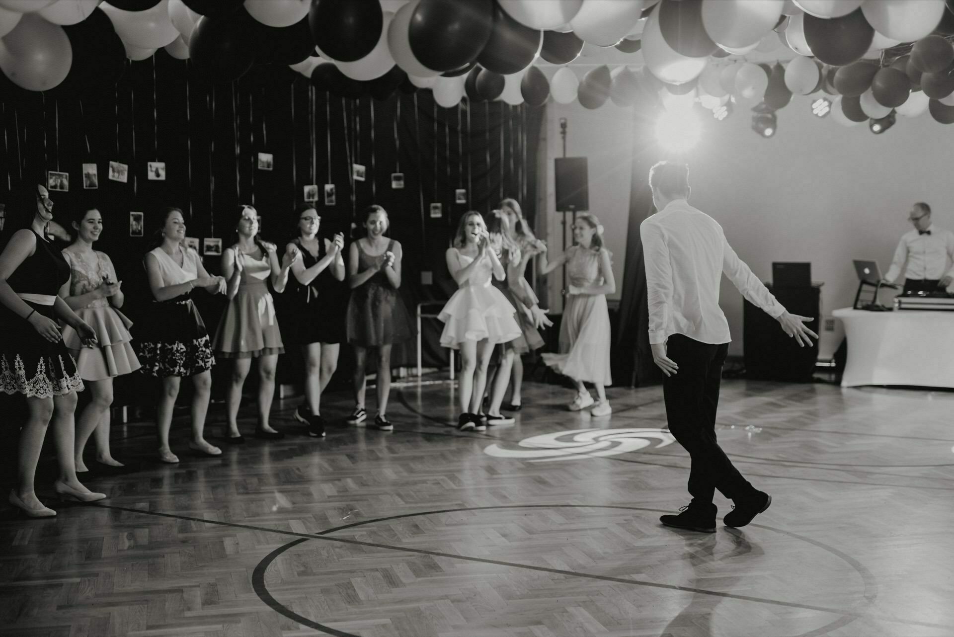 A person dances in the middle of a decorated room, while an elegantly dressed crowd claps and smiles from the sidelines. Balloons hang from the ceiling, illuminated by a spotlight. In the background, a DJ stands at a booth and captures the perfect moments for event photojournalism by a talented event photographer Warsaw.  