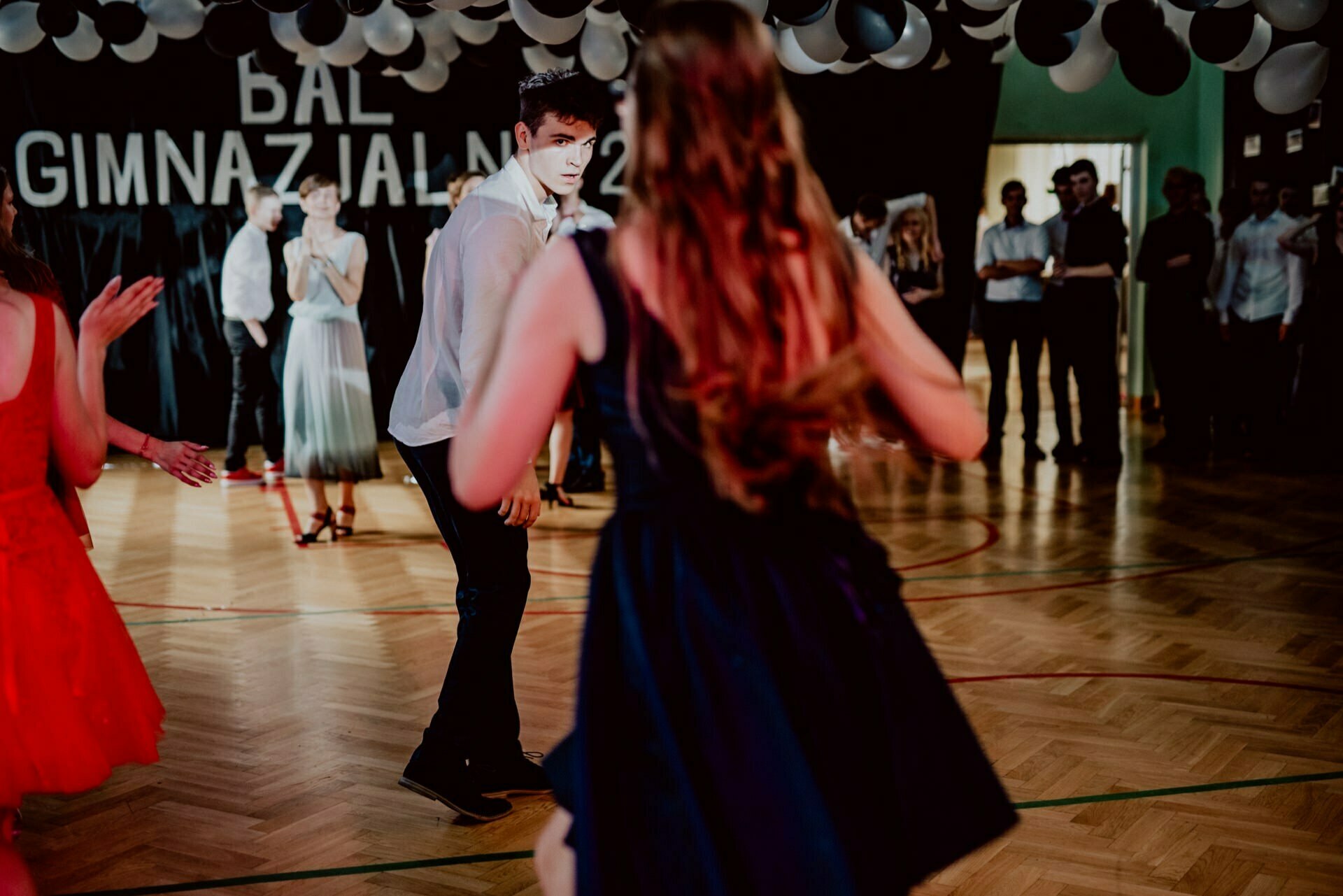 A group of people dancing in a decorated room with a wooden floor. A person in a white shirt turns toward a person in a dark dress in the foreground. At the top, black and white balloons form an arch, and the wall reads "Gymnasium Ball." The scene was beautifully captured by event photography.   