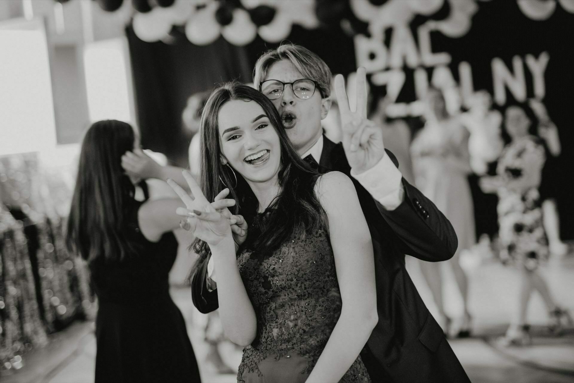 The black-and-white photo shows a young woman and man smiling and playfully posing. The woman in front, wearing a sleeveless dress, holds a peace sign, while the man behind her in glasses and a suit makes a peace sign next to her head. This is perfect event photography with people and balloons in the background.  