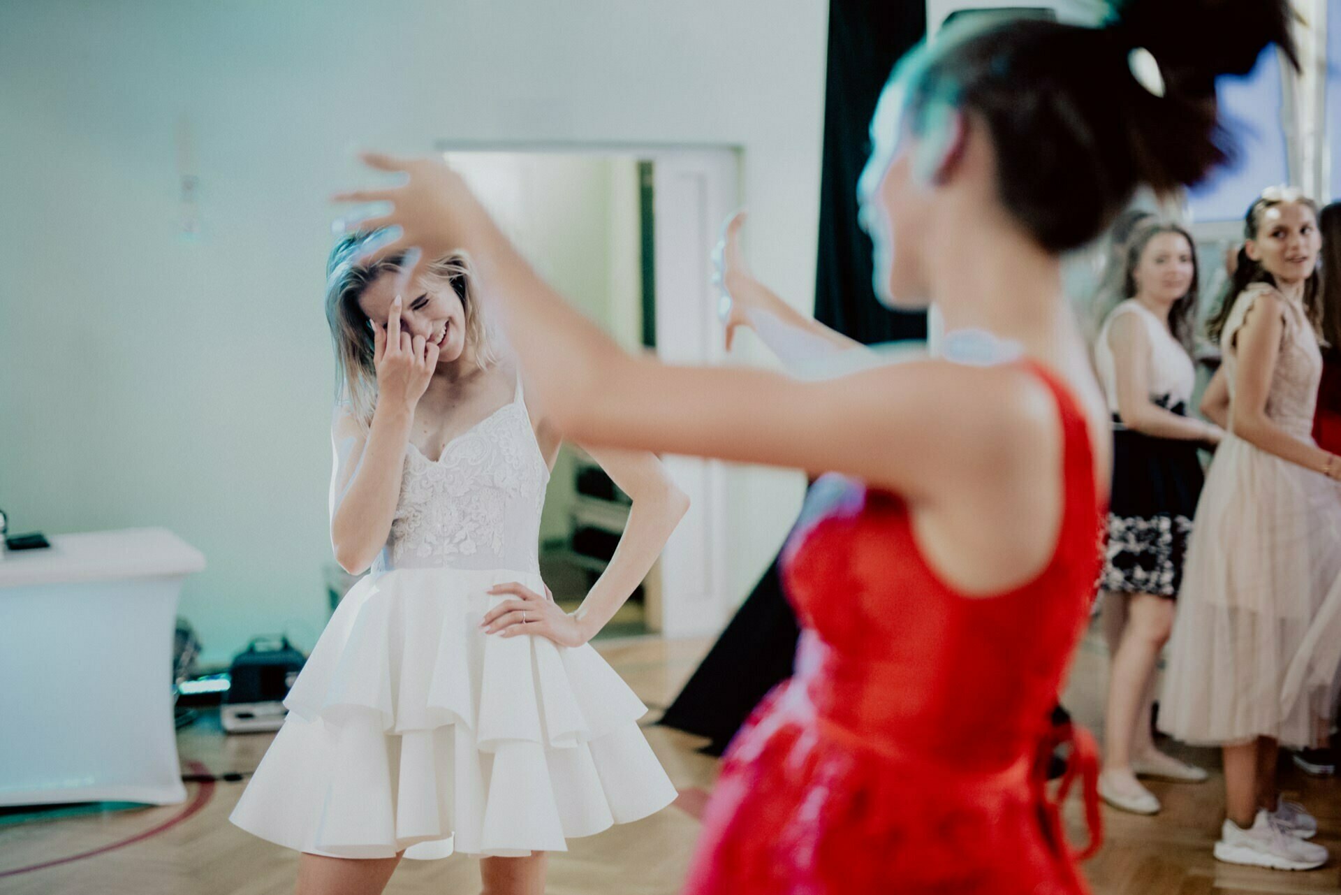 A woman in a white dress smiles, covering her face with her hand, while another woman in a red dress dances vigorously in the foreground. Several people in the background look on and smile. The scene, beautifully captured as event photography, appears to be a joyful party in a room.  