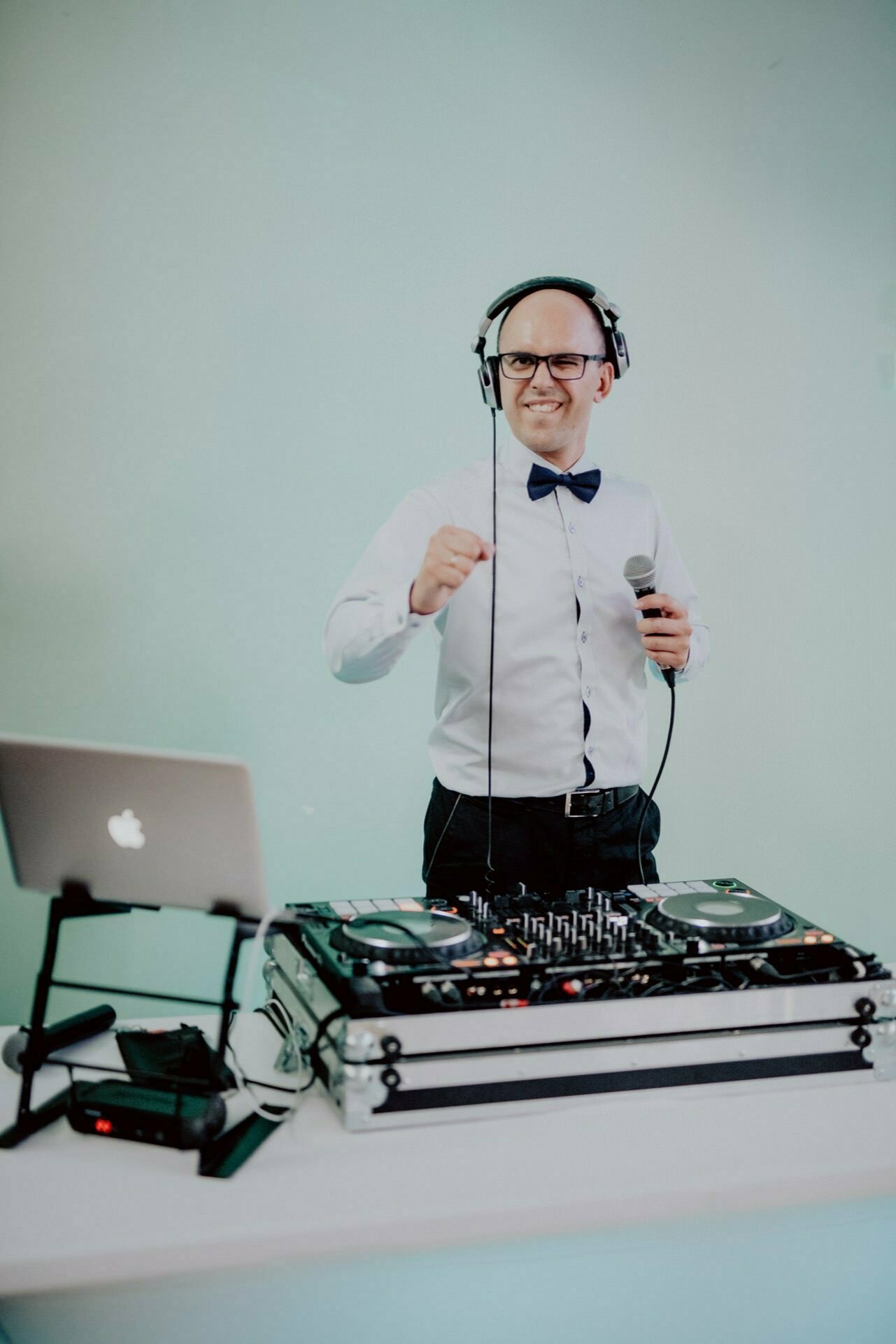 Behind the DJ equipment stands a bald man wearing glasses, a white shirt, dark pants and a bow tie. He is smiling, wearing headphones and holding a microphone. To his left is a laptop and in front of him is audio equipment against a pale green wall - perfect for event photography.  